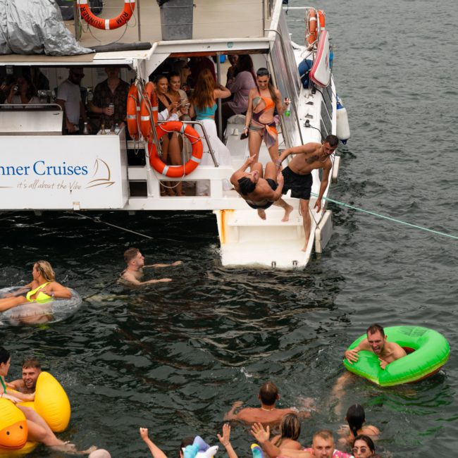 People are enjoying a Catamaran party in Sydney; some swim with pool floats while others stand onboard. A man is seen diving into the water from the boat's stairway.
