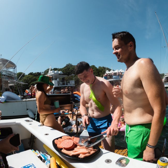 Two men in swim trunks are grilling burgers on a boat during a lively Sydney boat party hire. A woman in a bikini stands nearby holding a drink, with other boats and people visible in the background.