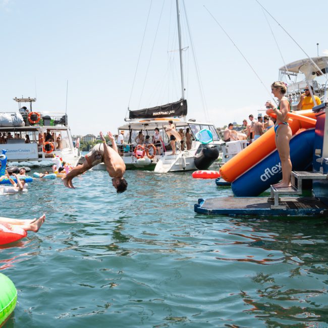 A group of people are enjoying a sunny day on the water with various boats and inflatables. A person is captured in mid-air performing a backflip into the water during a lively Sydney boat party hire event.