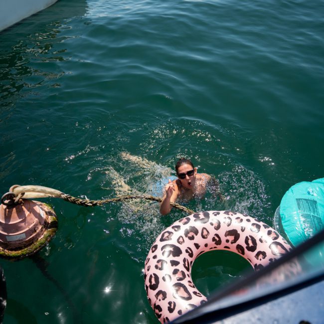 A person is swimming near a boat, holding onto an inflatable leopard print inner tube while another person is seated on the boat. The water is calm and clear, and a buoy is visible, all set against the scenic backdrop of a luxury yacht hire in Sydney.