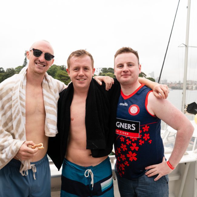 Three men stand on a boat, smiling at the camera. Two have towels draped over their shoulders, and one wears a sleeveless shirt with various graphics. A waterfront park is visible in the background, highlighting the beauty of a private yacht charter on Sydney Harbour.