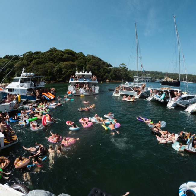 A group of people on inflatable rafts and floating toys enjoy a sunny day on the water, surrounded by several anchored boats. Trees and a shoreline are visible in the background, enhancing this scenic moment reminiscent of a luxury yacht hire in Sydney.