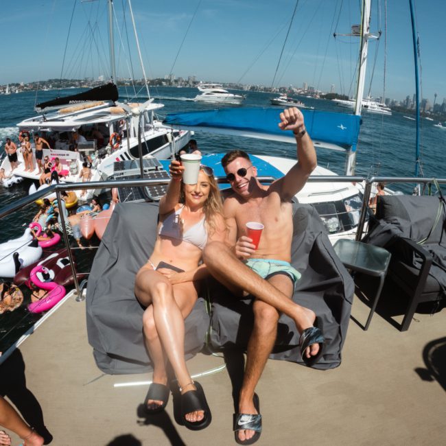 Two people sitting on a boat raise their drinks. Other boats and people are visible on the water in the background. The city skyline is visible in the distance, capturing the perfect setting for a Catamaran party Sydney.