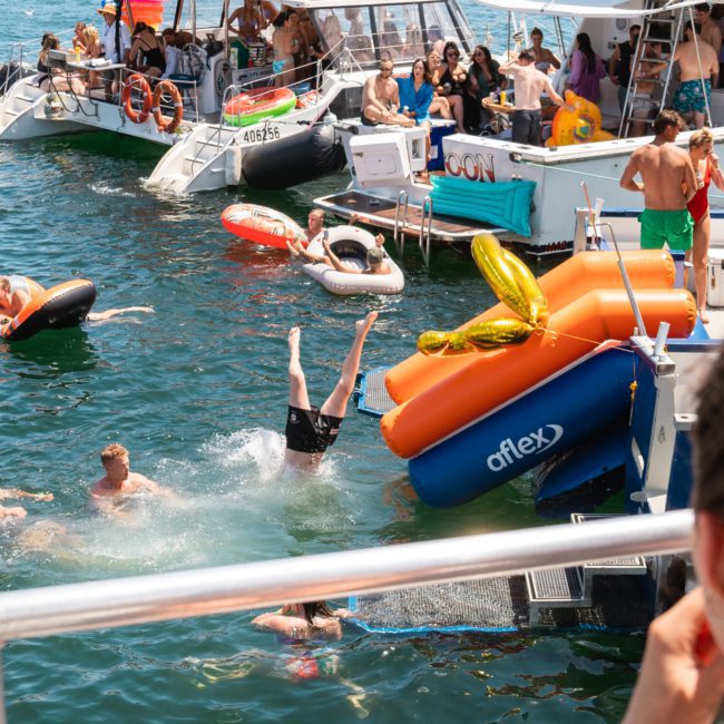 People enjoy swimming and lounging on inflatables in the water between boats during a lively summer gathering. A person is mid-dive off one of the boats, setting the perfect scene at a luxury yacht hire Sydney event.