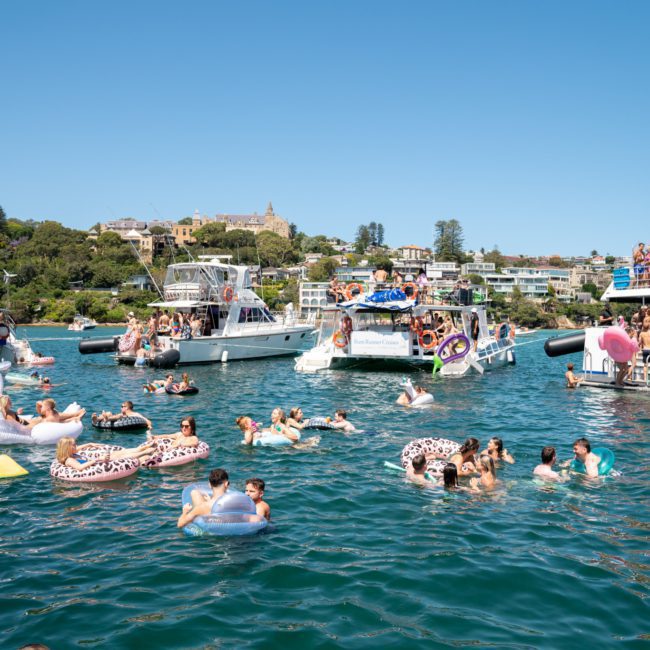 A lively scene of people enjoying a sunny day on the water, with many on boats and others on inflatables. The shoreline in the background has buildings and trees, perfect for a Sydney boat party hire or corporate boat events Sydney.