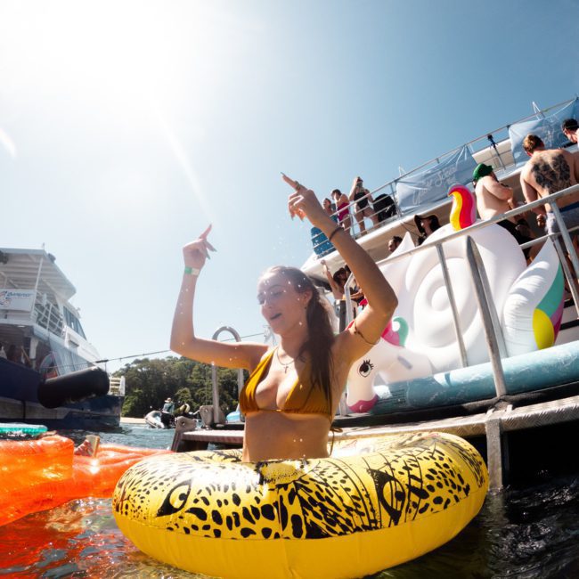 Woman in a yellow inflatable ring enjoys a sunny day in the water, with other people and floats in the background near a dock. Perfect setting for a private yacht charter Sydney Harbour.
