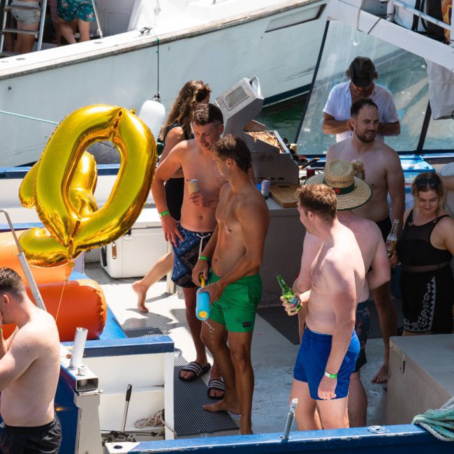 People in swimwear gather on a boat, holding a large gold balloon shaped like a number zero. Various individuals are interacting, some holding drinks, with an orange slide visible on the left. It's the perfect scene for Sydney boat party hire or a stylish luxury yacht hire Sydney event.