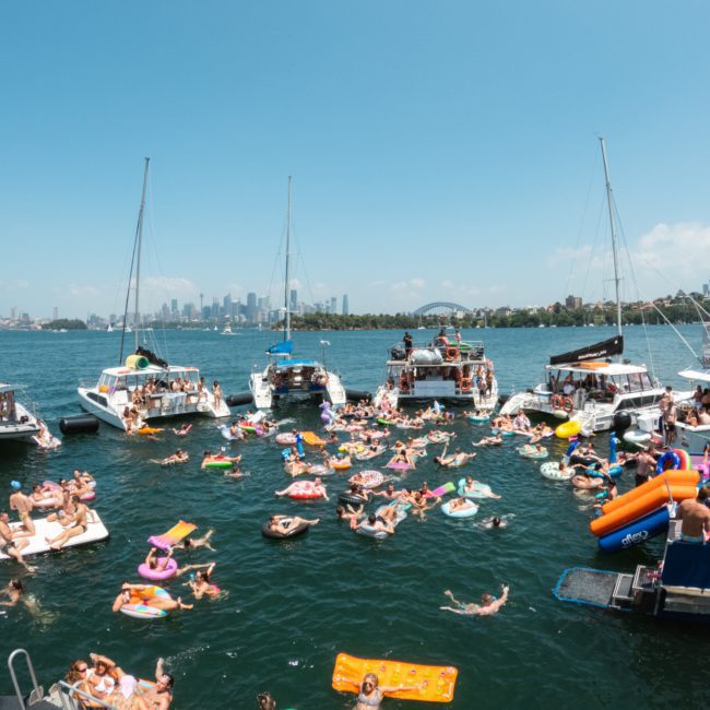 A large group of people on inflatables and swimming by anchored boats in a sunny bay with a city skyline in the background, enjoying the vibrant atmosphere of a Sydney boat party hire.