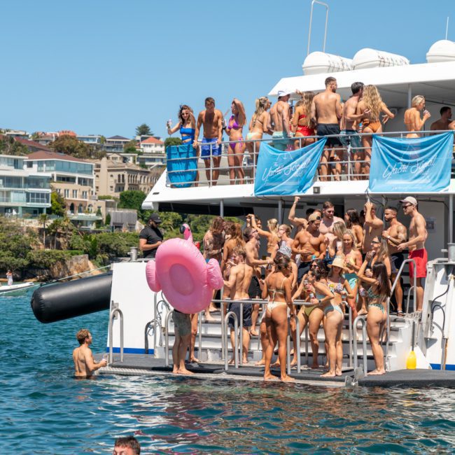 A lively corporate boat event in Sydney with people in swimwear enjoying the sun. Some individuals are on the lower deck in the water while others gather on the upper deck. Buildings and greenery are visible in the background, creating a perfect atmosphere for a memorable DJ boat hire Sydney experience.