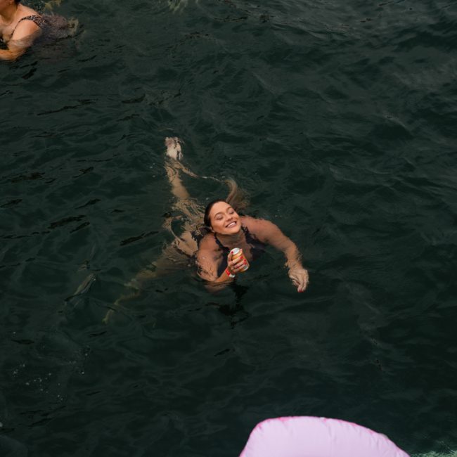 A person is swimming in dark water, holding a can and smiling, with others nearby relaxing on flotation devices during a luxury yacht hire Sydney.