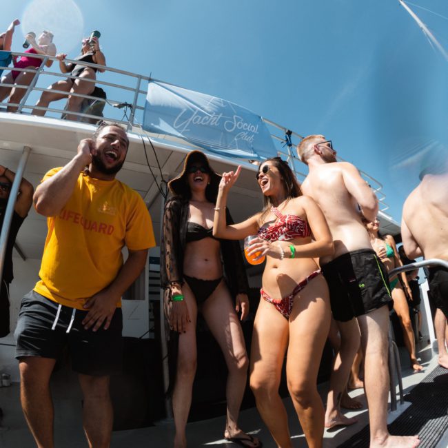 A group of people in swimwear are enjoying themselves on a boat. The person in a yellow shirt laughs with others under a bright blue sky. The boat, perfect for a private yacht charter on Sydney Harbour, has a sign that reads "Yacht Social Club.