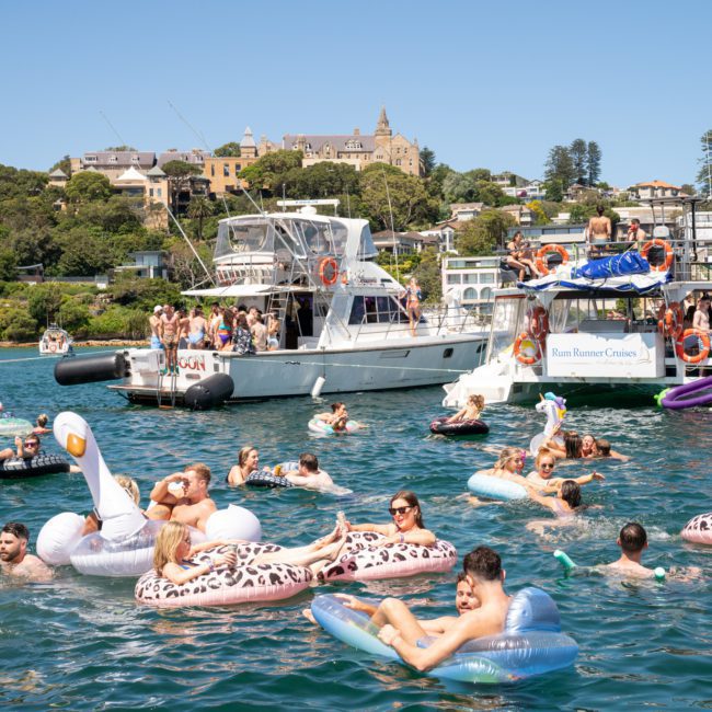 People are swimming and relaxing on inflatable floats in a bay, surrounded by anchored luxury yachts, with a backdrop of houses and greenery under a clear blue sky.