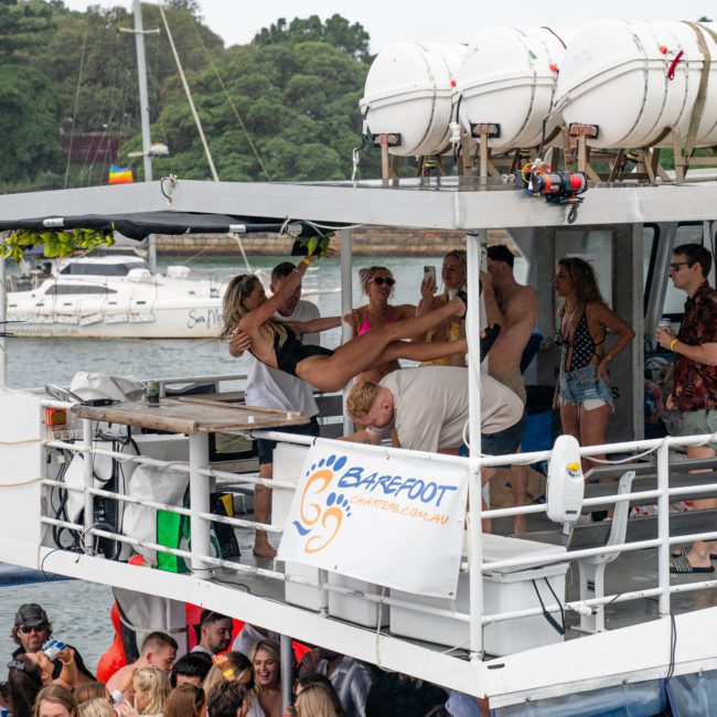 Partygoers are dancing and celebrating on a boat's deck adorned with a "Barefoot Downunder" banner, with more boats and a forested shore visible in the background—a perfect snapshot of Sydney boat party hire.