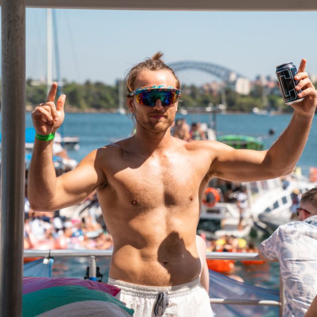 A shirtless man wearing sunglasses, holding a beverage, and posing with both hands raised on a catamaran party in Sydney. The background includes water and a bridge.
