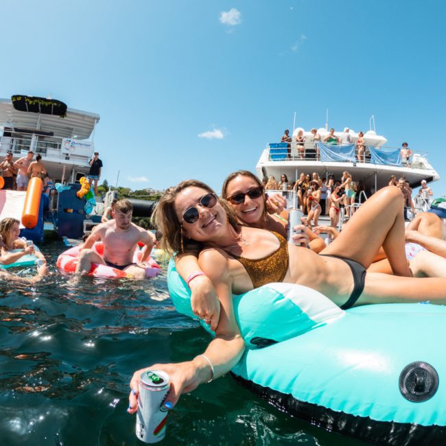 People enjoying a sunny day on a lake with floating inflatables, drinks in hand, and a luxury yacht hire Sydney in the background.