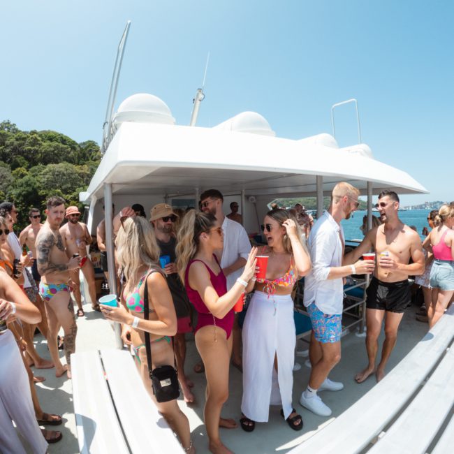 A group of people in swimwear socializes and drinks on a luxury yacht hire Sydney under a clear blue sky. Various conversations and activities are taking place on the deck. Trees are visible in the background.