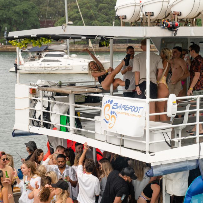 A group of people dancing and socializing on a two-story boat named "Barefoot Charters." The scene shows both levels of the boat with attendees in swimwear, a nearby marina, trees in the background, and lively music from a DJ—perfect for Corporate boat events Sydney.