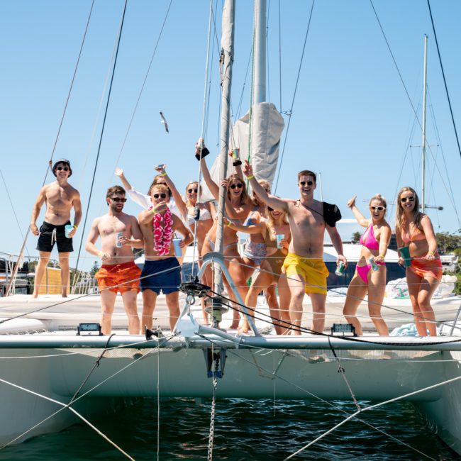 A group of people in swimsuits stands joyfully on the deck of a catamaran party in Sydney under a clear sky, raising their arms and cheering, with other boats visible in the background.