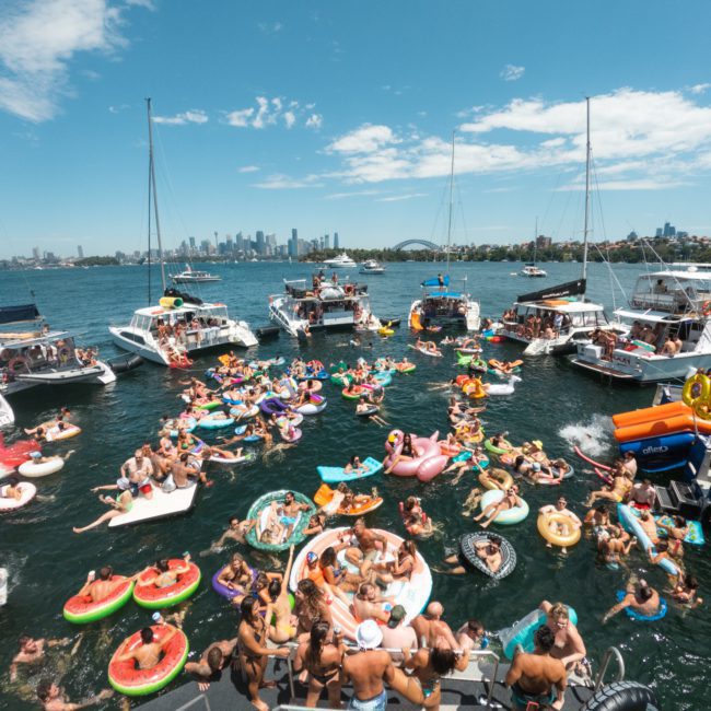 A crowded gathering of people on inflatable floaties in the water, surrounded by several anchored yachts and a catamaran party Sydney, with a city skyline visible in the background under a clear blue sky.