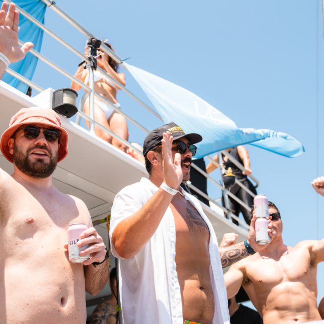 A group of individuals stands on a boat, holding drinks and raising their hands; some are wearing hats and sunglasses, while others have visible tattoos. Sydney boat party hire offers an unforgettable experience on the water.
