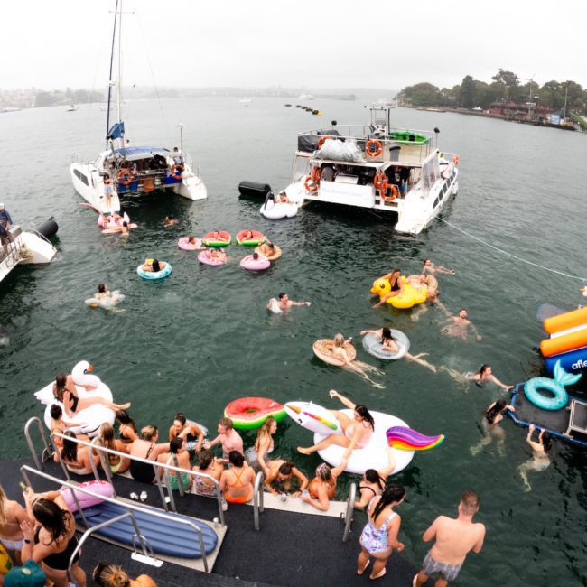 People are swimming and floating on colorful inflatables in a body of water near docked boats, enjoying a lively party. Multiple boats, including luxury yacht hires in Sydney, have gathered in close proximity under the cloudy sky.