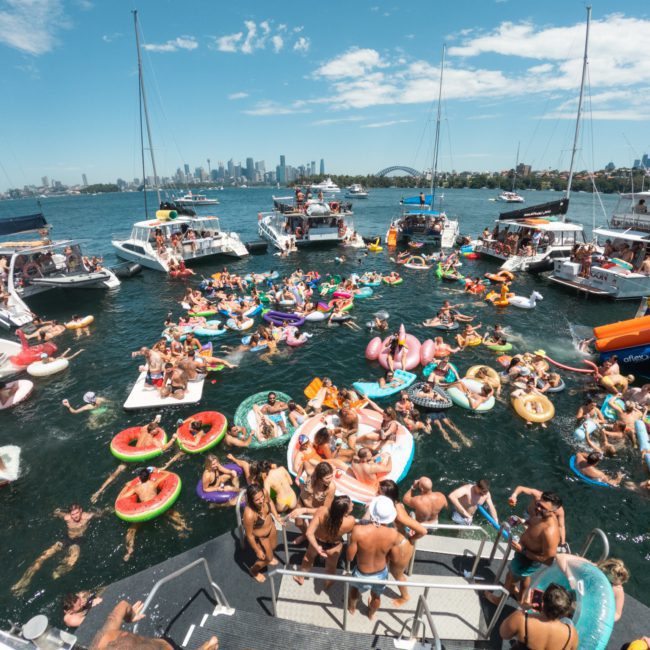 A large group of people are enjoying a sunny day on the water, surrounded by boats and floating on inflatable rafts. The city's skyline is visible in the background, creating the perfect setting for a Sydney boat party hire or corporate boat events Sydney.