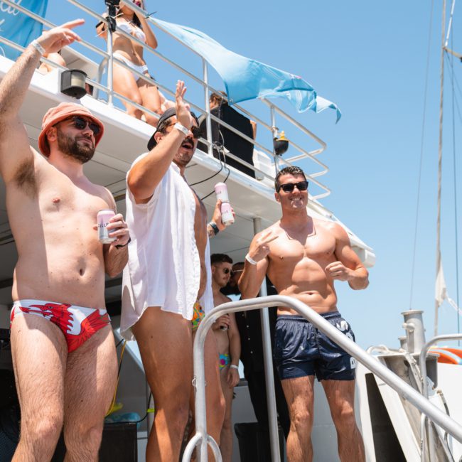 Three men in swimwear are standing on a catamaran's deck, each holding a drink. Two are shirtless, and one wears a sleeveless white shirt. Other people can be seen in the background enjoying the Sydney boat party hire atmosphere.