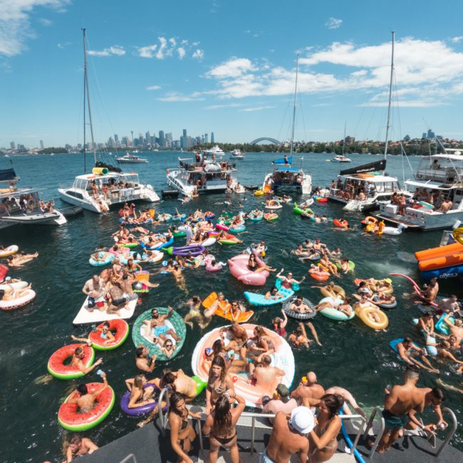 Numerous people are floating on colorful inflatables and boats, socializing and enjoying a sunny day on a bustling waterway with a city skyline visible in the background, reminiscent of lively corporate boat events in Sydney.