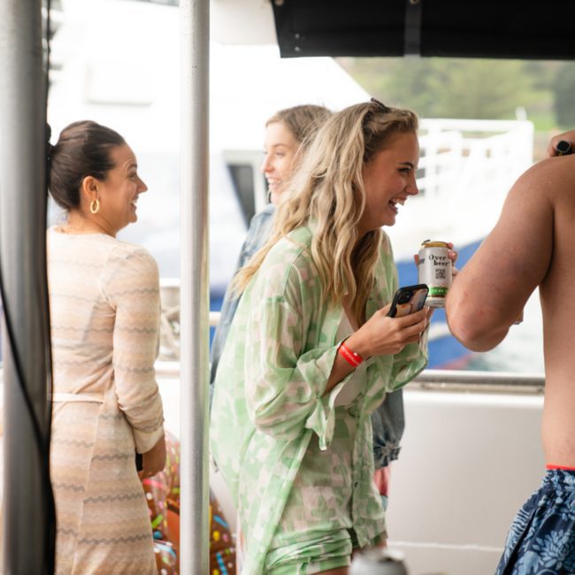 Two women and a man interact and smile on a boat during a lively Catamaran party in Sydney, with one woman holding a canned drink and phone.
