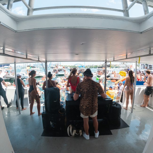 Rear view of a DJ performing on a boat with a crowd of people in swimwear on the deck and in the water. Various boats, including catamarans, are visible in the background as part of an unforgettable Catamaran party Sydney experience.