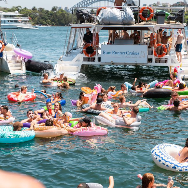 A large group of people on inflatable floats enjoy a sunny day on the water near a docked boat labeled "Rum Runner Cruises." In the background, a bridge is visible, setting an ideal scene for a Catamaran party in Sydney.