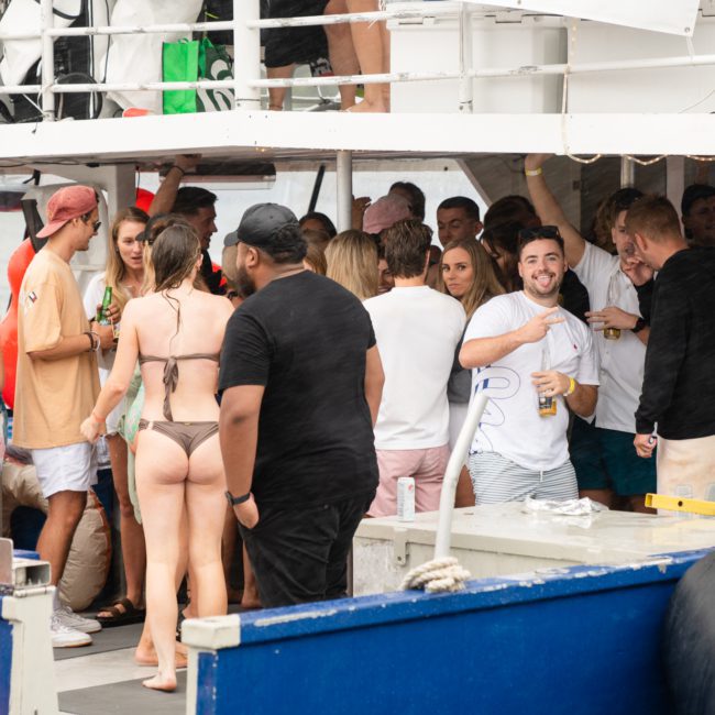 A group of people socializing on a luxury yacht hire in Sydney; some are standing and chatting, while others are holding drinks. The attendees are dressed in casual summer attire, with swimwear visible.