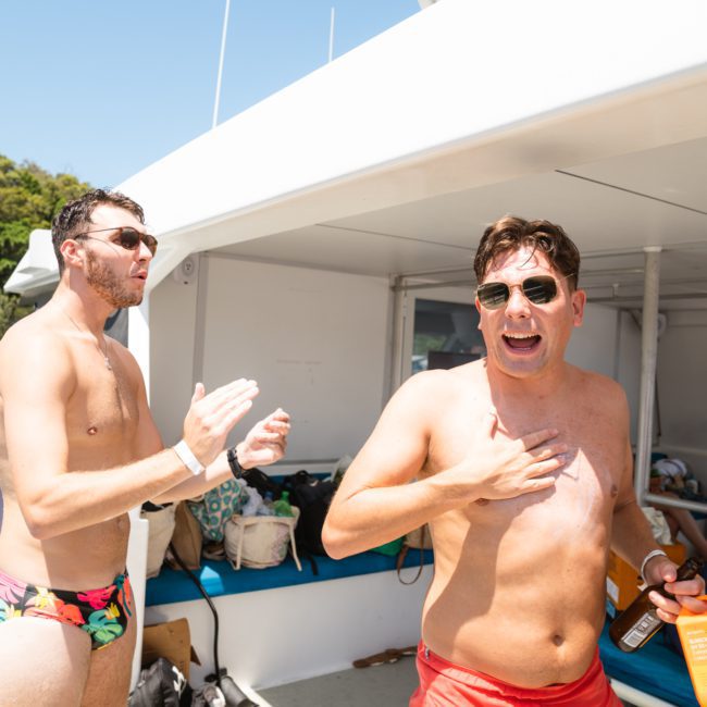 Two men in swimwear are on a luxury yacht under sunny weather. One man is applying sunscreen to his chest while the other stands nearby, clapping his hands.