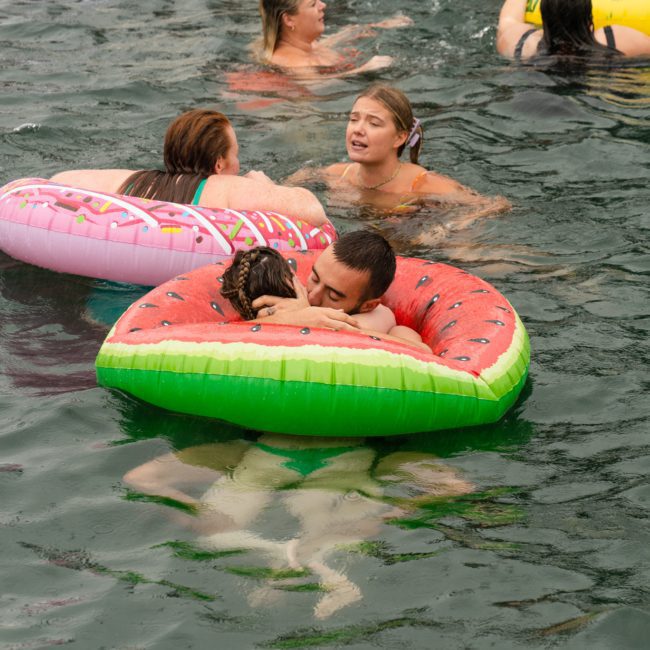 People swimming in a lake, with some individuals floating on inflatable rings shaped like a watermelon slice and a donut. One person appears to be kissing a child on the watermelon float, while others chat about planning their next luxury yacht hire Sydney event.