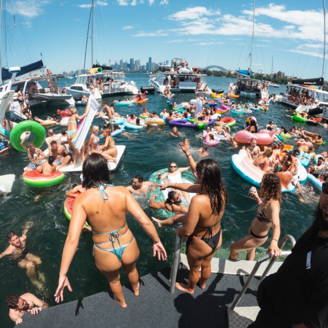 A large group of people enjoying a party on a lake, with floaties and boats in the background. People are swimming, socializing, and sunbathing under a blue sky. Highlights include DJ boat hire Sydney, adding an electrifying vibe to the celebration.