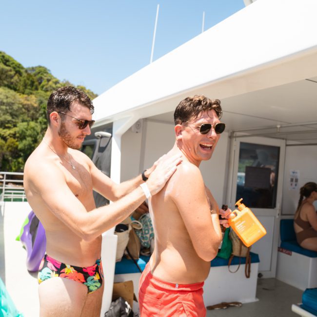 Two men in swimwear on a private yacht charter in Sydney Harbour; one applies sunscreen to the other's back. Background includes a boat interior, several seated people, and lush greenery.