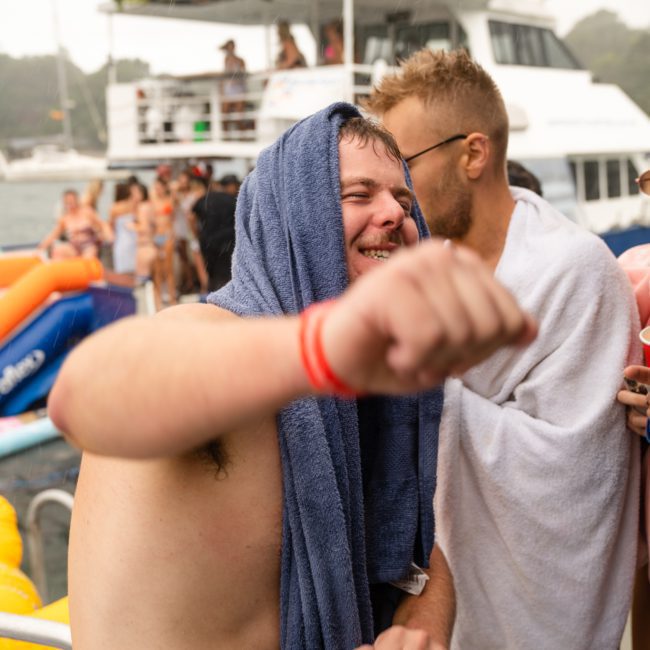 Two people wrapped in towels appear to be enjoying themselves on a private yacht charter in Sydney Harbour, with a crowd and another boat visible in the background. One person is mid-motion, possibly dancing.