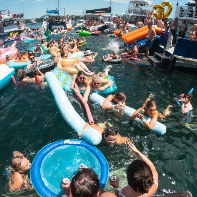 People in swimsuits enjoying a lively pool party, floating on inflatables and swimming near luxurious yachts.