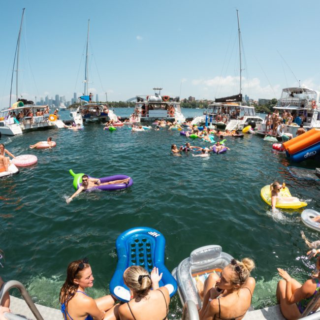 A crowd of people are enjoying a sunny day on a lake, floating on inflatable rafts and surrounded by multiple anchored boats, reminiscent of a Sydney boat party hire.