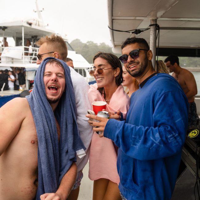 A group of people, some in swimwear, are gathered on a boat. One person has a towel draped over their shoulders, and another is wearing sunglasses and holding a red cup. Other boats are in the background, adding to the festive atmosphere of this Sydney boat party hire.