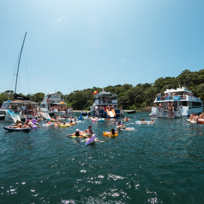 A group of people enjoying a sunny day in the water with boats anchored nearby, while others float on inflatable rafts and slides, creating a lively scene perfect for a luxury yacht hire Sydney experience.
