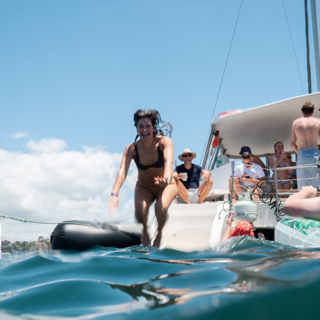 A woman jumps into the water from a ladder on a docked boat while several other people lounge and relax on the deck of a luxury yacht. The sky is clear and sunny, perfect for a private yacht charter in Sydney Harbour.