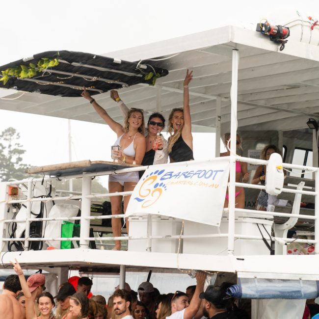 A group of people on a boat, with some on the upper deck holding drinks and smiling. A banner that says "Barefoot" is hanging on the railing. There are trees and water in the background, making it a perfect scene for a Catamaran party Sydney style.