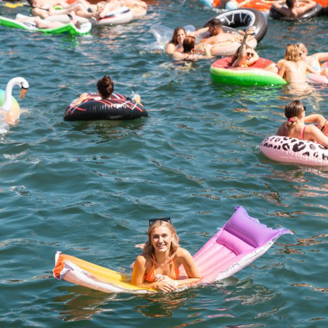 People floating on various inflatables in the water at a crowded outdoor event on a sunny day. A woman is smiling on a colorful inflatable near the center, reminiscent of a *catamaran party Sydney* experience.
