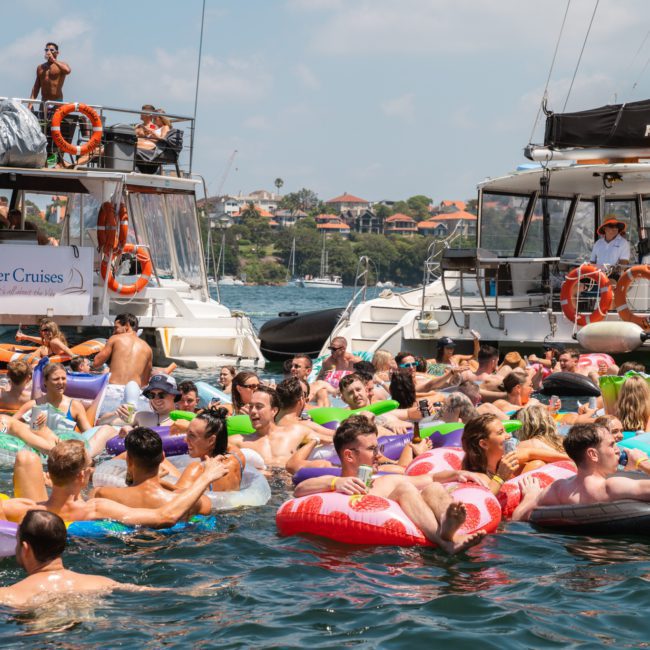 A group of people in inflatable floaties enjoying a sunny day on the water, surrounded by two boats labeled "Rum Runner Cruises" and "RockfishMV," reveling in the fun atmosphere of a catamaran party in Sydney.