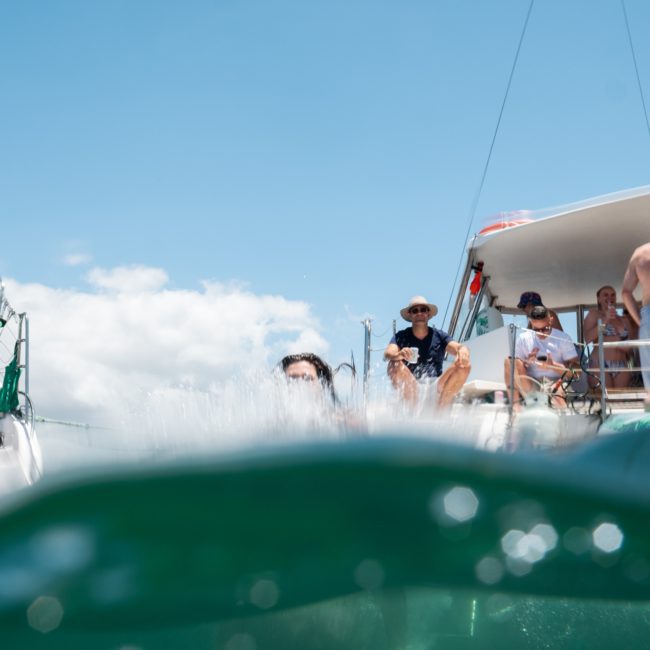 People on a boat enjoying a sunny day with some individuals swimming in the water. Boats are anchored nearby, perfect for a Sydney boat party hire. The water splashes in the foreground, adding to the lively atmosphere.
