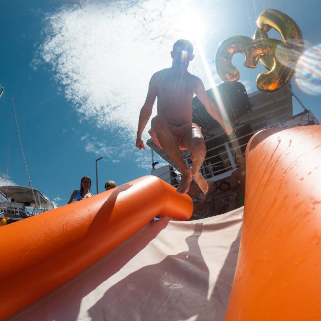 A person in swim trunks is jumping off an inflatable orange slide with sunglasses on, under a bright sunny sky during a Sydney boat party hire. A golden balloon shaped like the number 33 is visible in the background.
