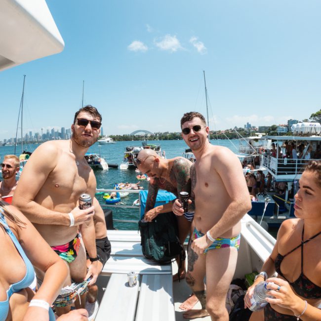 Group of people in swimwear enjoying a party on a luxury yacht hire Sydney on a sunny day. Some hold drinks while others socialize. Boats and city skyline in the background, perfect for those looking into DJ boat hire Sydney.