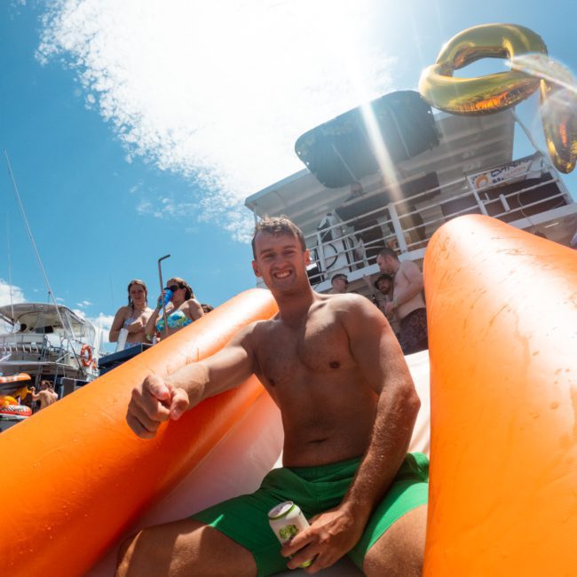 A man in green shorts sits on an orange inflatable slide holding a drink, with a boat and clear sky in the background. Other people are visible in the distance, adding to the lively atmosphere of luxury yacht hire Sydney.