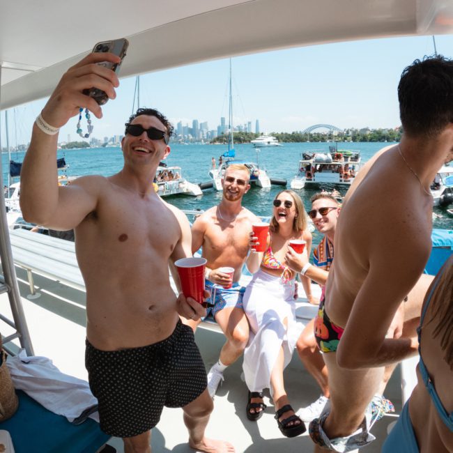 A group of people on a boat taking a selfie, holding red cups. The ocean and other boats are in the background on a sunny day, showcasing the excitement of corporate boat events Sydney.